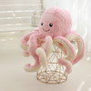 Pink baby octopus plush toy with smiley face sitting on a wire basket, ideal for kids' playroom decor and cuddling.