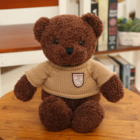 Brown teddy bear plush toy wearing a beige sweater, sitting on a wooden table for decoration or gift purposes.