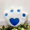 White plush pillow with blue heart and paw print design, surrounded by green leaves and white flowers.