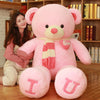 Large pink teddy bear with heart details, suitable as a plush gift for Valentine's Day, beside a smiling woman in a cozy room.