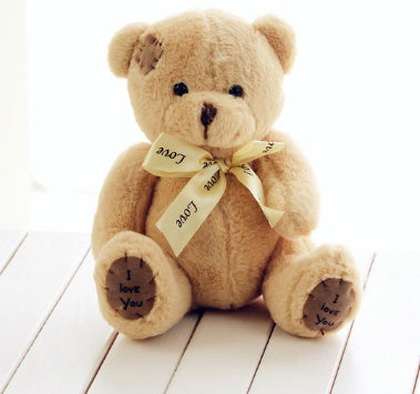 Cute patch teddy bear plush with "I love you" foot pads and a "Love" ribbon, sitting on a white wooden surface.