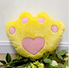 Soft yellow paw-shaped plush pillow with pink heart and paw prints, surrounded by green leaves and flowers.