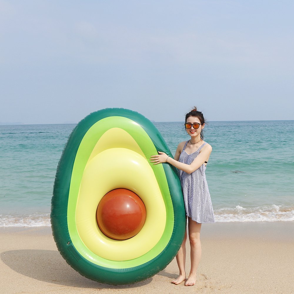 Woman holding a giant inflatable avocado pool float on a sandy beach with the ocean in the background.