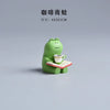 Green frog figurine holding a coffee cup and book; dimensions 4x3x2cm.