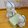 Two plush dolls in white protective suits playing with a green box in a colorful room.