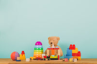 Colorful toys including blocks, cars, and a teddy bear on a wooden table with a light blue background.