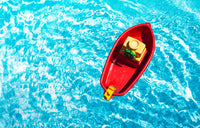 Small red toy boat floating on clear blue water in a swimming pool.