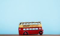 Vintage red toy double-decker bus with UNLOP logo on a wooden surface, set against a bright blue background.